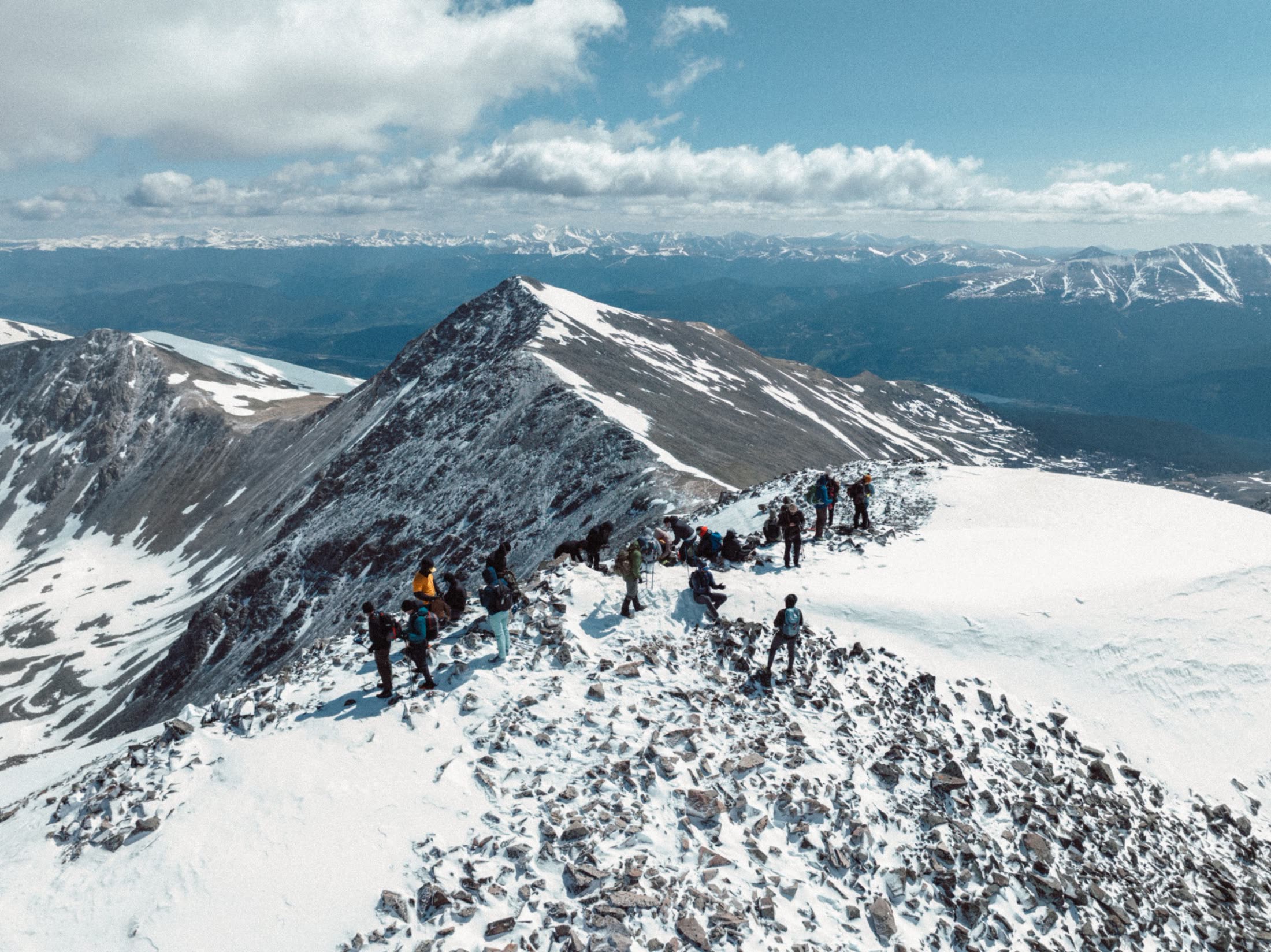 Climbers resting on a snowy summit ridge above a sharp peak and distant ranges