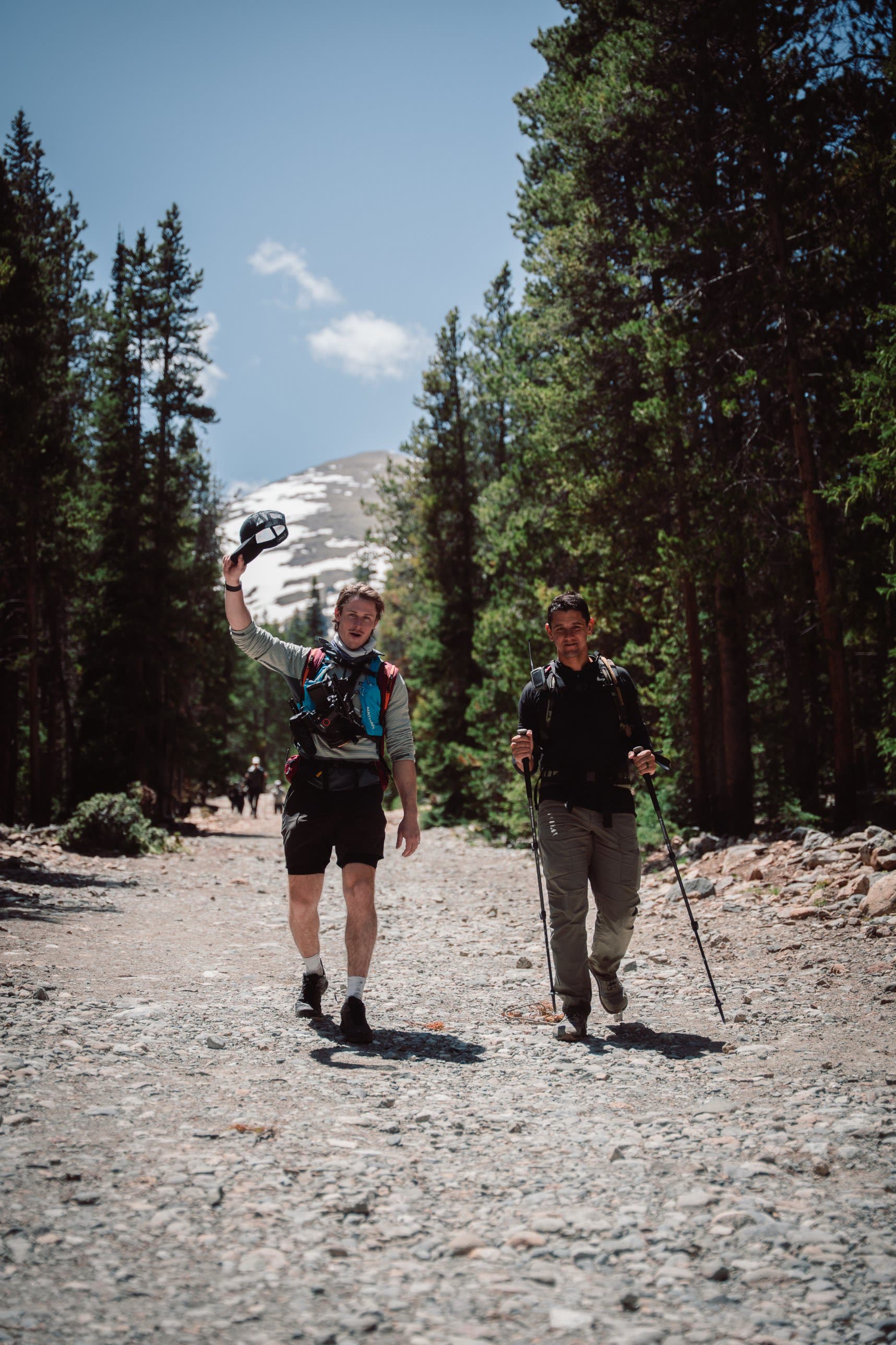 Two hikers walking a forest trail toward the camera, one waving his cap