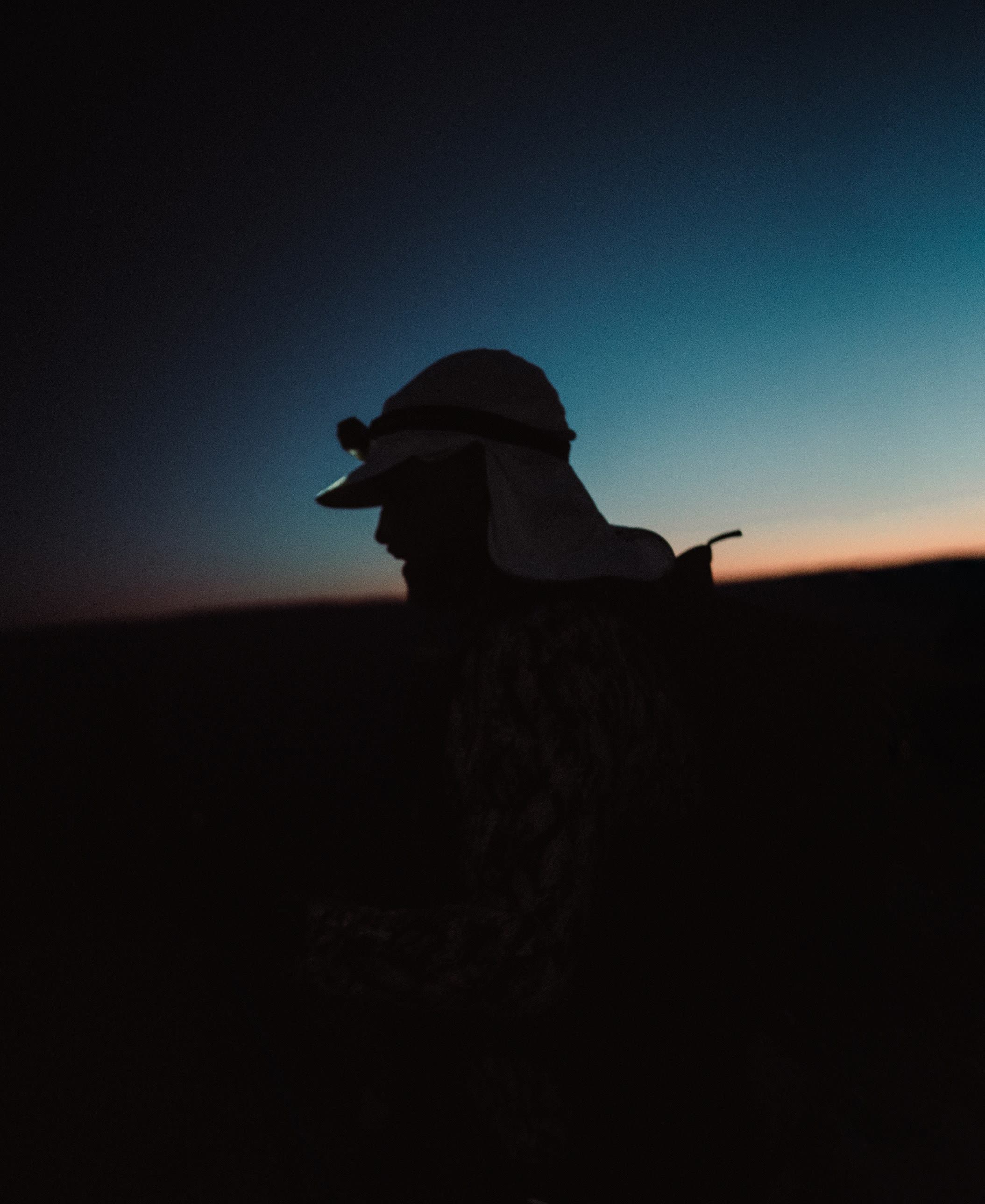 A hiker in a headlamp and sun hat silhouetted against the predawn sky